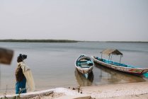 Yoff, Senegal - December 6, 2017: Side view of woman walking on shore of river with shabby sailboats in sunlight . — стоковое фото
