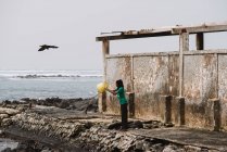 Goree, Senegal- Dezembro 6, 2017: Vista lateral da mulher de pé perto do barracão em ruínas e derramando água suja para o mar . — Fotografia de Stock