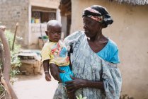 Yoff, Senegal- 6 de diciembre de 2017: Mujer anciana sosteniendo a un niño pequeño en las manos sobre el fondo de la aldea rural . - foto de stock