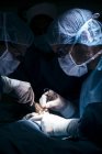 Vista lateral de dois cirurgiões operando paciente na luz da lâmpada — Fotografia de Stock