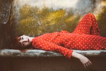 Mulher ruiva em roupas vermelhas deitada no banco de pedra musgosa e olhando para a câmera — Fotografia de Stock