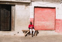 Goree, Senegal- 6 dicembre 2017: Bambino africano a piedi nudi seduto sulla panchina in strada asfaltata in piena luce . — Foto stock