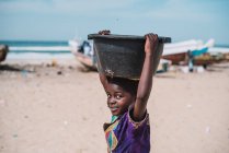 Гори, Сенегал - 6 декабря 2017 года: Портрет девушки, несущей пластиковый бассейн на голове и смотрящей в камеру на пляже — стоковое фото