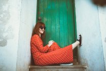 Femme rousse en robe rouge et lunettes de soleil assis sur le seuil de la porte — Photo de stock