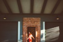 Stylische rothaarige Frau in roter Kreisel gegen Tür im Sonnenlicht. — Stockfoto