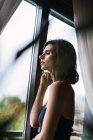 Vista laterale della donna bruna con trucco luminoso in piedi vicino alla finestra e guardando fuori — Foto stock