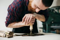 Bärtiger professioneller Handwerker, der Goldschmuck auf ein Stück Leder prägt. — Stockfoto