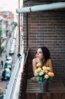 Menina morena sentada à mesa com flores em vaso na varanda e olhando para longe — Fotografia de Stock
