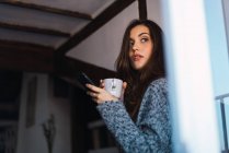 Брюнетка дівчина з чашкою кави і смартфон в руках, що хтось дивитися вбік — стокове фото