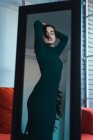 Portrait sensual brunette in green dress standing in front of mirror and posing with arms raised and eyes closed. — Stock Photo