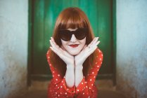 Portrait of redhead woman in red clothing and sunglasses posing with chin on hands — Stock Photo