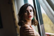 Brunette girl with red lips hugging shoulder and posing near window — Stock Photo