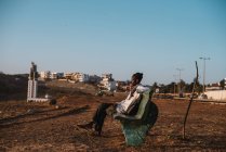 Goree, Senegal- 6 de diciembre de 2017: Vista lateral del hombre sentado en el banco en el campo seco y sucio en el fondo de la pequeña ciudad . - foto de stock