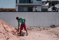 Goree, Senegal- 6 de diciembre de 2017: Trabajador negro bajo la luz del sol cargando arena en cubo con pala . - foto de stock