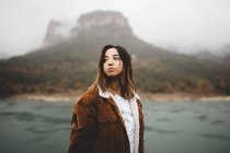 Pretty young woman standing near river on background of hill. — Stock Photo