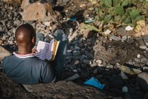 Über dem Kopf eines afrikanischen Mannes, der Buch liest, während er auf Felsen an der Küste sitzt. — Stockfoto