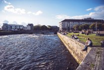 GALWAY, IRLANDA - 9 de agosto de 2017: Las personas que tienen tiempo en el césped iluminado por el sol cerca del canal - foto de stock