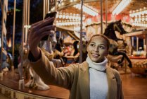 Smiling woman in coat standing near merry-go-round and taking selfie. — Stock Photo