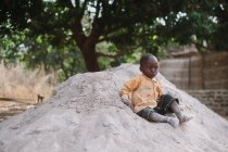 Kedougou, Senegal- 6 de diciembre de 2017: Retrato de un niño sentado en un montón de arena en un pueblo rural y mirando seriamente hacia otro lado . - foto de stock