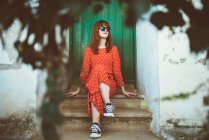 Redhead girl in sunglasses sitting on doorstep and looking away — Stock Photo