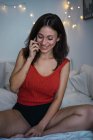 Smiling brunette girl sitting on bed and talking on smartphone — Stock Photo