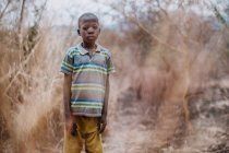 Goree, Senegal- diciembre 6, 2017: Niño negro con ropa sucia parado en el campo seco rural y mirando a la cámara  . - foto de stock