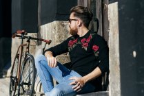 Portrait of man in sunglasses sitting near bicycle on street and looking away — Stock Photo