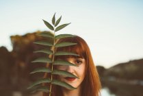 Portrait of ginger woman with red lips posing playfully and covering eye with leaves — Stock Photo