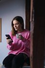 Случайная женщина в розовом фотке пьет кофе и просматривает смартфон дома — стоковое фото