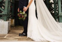 Vista lateral da colheita do noivo com noiva elegante posando juntos e de mãos dadas . — Fotografia de Stock