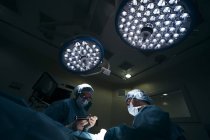 Vista de ángulo alto de las lámparas bajo los cirujanos en uniforme teniendo cuidado del paciente en quirófano - foto de stock