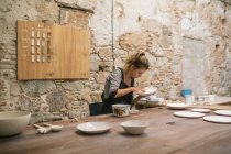 Концентрована жінка в фартусі сидить за столом і формує тарілки з білої глини . — стокове фото