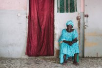 Goree, Senegal- Dezembro 6, 2017: Mulher sênior em azul sentado perto da porta da casa e olhando para a câmera . — Fotografia de Stock