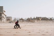 Yoff, Senegal- 6 de diciembre de 2017: Vista lateral del niño negro relajándose en la playa tropical de arena del océano con sombrillas de paja en el fondo . - foto de stock