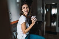 Усміхнена брюнетка дівчина сидить за кухонним баром і п'є каву — стокове фото
