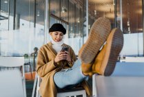 Молодая женщина в теплой одежде с ногами на столе и с помощью смартфона, сидя в кафе . — стоковое фото
