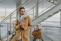 Portrait of young woman wrapping in coat and leaning on bicycle in front of staircase. — Stock Photo