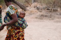 Niokolo koba, Sénégal- 8 décembre 2017 : Vue de côté de la femme africaine en vêtements brillants portant un nouveau-né derrière le dos dans un village rural . — Photo de stock