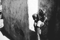 Goree, senegal- 6. Dezember 2017: Teenager mit kleinem Baby auf dem Rücken steht im Sonnenlicht in der Nähe eines Dorfgebäudes. — Stockfoto
