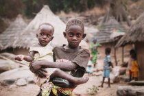 Yoff, Sénégal- 6 décembre 2017 : Un petit Africain tenant une charmante petite sœur sur les mains et regardant une caméra dans un village rural . — Photo de stock
