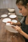 Mulher concentrada sentada à mesa e criando pratos de barro branco . — Fotografia de Stock
