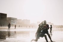 Yoff, Sénégal- 6 décembre 2017 : Des jeunes hommes luttent et rient dans des éclaboussures de vagues océaniques sur la plage . — Photo de stock