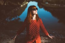 Young redhead woman in red clothing and sunglasses posing confidently in sunlight over lake. — Stock Photo