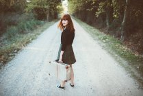Ginger woman holding suitcase and on rural road smiling at camera over shoulder. — Stock Photo