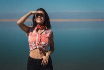 Brunette girl in summer outfit posing with palm at forehead on background of lake — Stock Photo