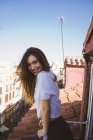 Вид збоку усміхненої брюнетки дівчина позує на балконі над дахами міський пейзаж і дивиться на камеру — стокове фото