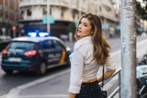 Brunette girl posing at street scene with blurred police car and looking over shoulder at camera — Stock Photo