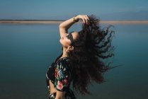 Вид збоку брюнетка дівчина в сонцезахисних окулярах махає волоссям над озерним пейзажем — стокове фото