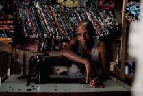 Goree, senegal- 6. Dezember 2017: Porträt eines Mannes, der sich in der Werkstatt an eine alte Nähmaschine lehnt und in die Kamera blickt. — Stockfoto