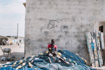 Goree, Senegal- December 6, 2017: Woman with child sitting on huge fishing net near concrete wall at street scene. — Stock Photo
